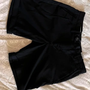 Svarta shorts med knappar - Svarta shorts med knappstängning och bälteshällor. De har en klassisk design med två bakfickor som stängs med knappar. 