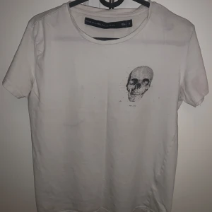 Vit t-shirt med dödskalletryck - Säljer en stilren vit t-shirt med ett coolt dödskalletryck på bröstet. Perfekt för en edgy look. T-shirten har en normal passform och är kortärmad. Skön och lätt att matcha med olika outfits.