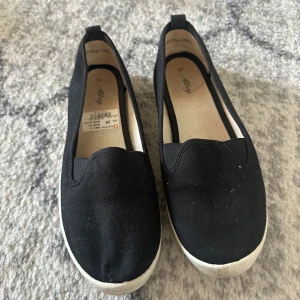 Svarta loafers från Alley - Säljer ett par svarta loafers från Alley. Skorna har en enkel design med vit sula och är perfekta för en avslappnad stil. De är lätta att ta på och av, vilket gör dem praktiska för vardagsbruk.