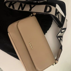 Beige axelväska från DKNY - Snygg beige axelväska från DKNY med justerbar axelrem i svart och beige med logotyp. Väskan har en elegant design med DKNY-logga framtill. Använd få gånger. Nypris 2500-3000kr