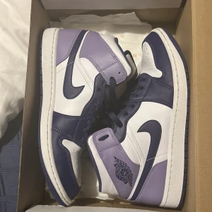 Nike Air Jordans i lila och vit - Snygga Nike Air Jordans i en fräsch kombination av lila och vit. Skorna har en klassisk hög design med snörning och den ikoniska Nike-loggan på sidan. Perfekta för att ge din stil ett lyft!