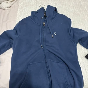 Blå hoodie från Polo Ralph Lauren - Säljer en blå hoodie från Polo Ralph Lauren med dragkedja och huva. Tröjan har en broderad logotyp på bröstet och praktiska fickor framtill. Perfekt för en avslappnad stil. Helt fräsch o ny, använd 1 gång pris kan diskuteras 