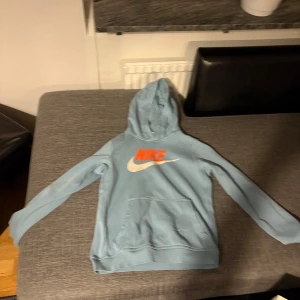 Ljusblå hoodie och byxor från Nike - Säljer en ljusblå hoodie och matchande byxor från Nike. Hoodien har en stor Nike-logga i orange och vitt på bröstet och en bekväm huva. Byxorna har samma logga på ena benet. Perfekt för en avslappnad stil.