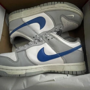 Nike Dunk i grått och blått - Snygga Nike Dunk sneakers i en stilren kombination av grått och vitt med en blå swoosh. Skorna har en klassisk design med snörning och en bekväm passform. Perfekta för en sportig look. Storlek 37,5. Använda 2-3 gånger men ser ut som nya. Därav priset. Tveka inte vid minsta fundering och skriv gärna😁