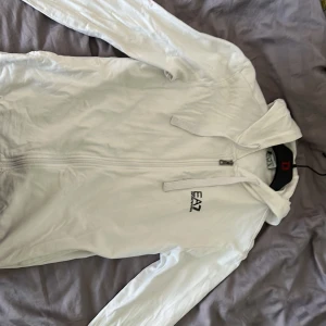 Vit huvtröja från EA7 - Säljer en stilren vit huvtröja från EA7 med dragkedja och huva. Perfekt för en avslappnad look. Den är rätt tunn så den e perfekt för en vår/sommar dag. 