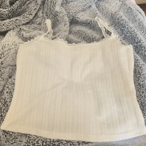 Vit ribbad linnetopp från Joocy Babi - Säljer en söt vit ribbad linnetopp från Joocy Babi. Toppen har spetsdetaljer vid axelbanden och en mjuk, bekväm passform. Perfekt för varma sommardagar eller som ett lager under en tröja.