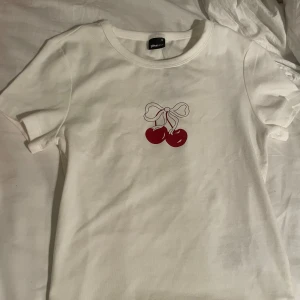 Vit t-shirt med körsbärstryck från Gina Tricot - Säljer en söt vit t-shirt från Gina Tricot med ett tryck av körsbär och en rosett på framsidan. T-shirten är kortärmad och har en normal passform. Perfekt för en avslappnad stil!