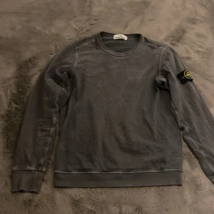Grå tröja från Stone Island - Säljer en stilren grå tröja från Stone Island med deras ikoniska märke på ärmen. Tröjan har långa ärmar och en rund halsringning. Köpt på NK i Stockholm.