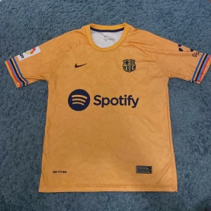 Gul FC Barcelona fotbollströja - Säljer en gul FC Barcelona fotbollströja från Nike med korta ärmar. Tröjan har klubbens emblem och sponsorlogga på framsidan samt 'Lewandowski 9' tryckt på baksidan. Ärmarna har färgglada ränder och tröjan är perfekt för alla Barça-fans! Den inkluderar små detaljer i tröjan, den är även i ett luftigt material perfekt för sommaren.