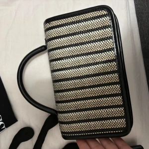 Randig handväska med svart och beige mönster - Snygg handväska med ett stilrent randigt mönster i svart och beige. Väskan har en praktisk handtag och är perfekt för att bära dina nödvändigheter. Den har en elegant design som passar till många olika tillfällen. Aldrig använd 