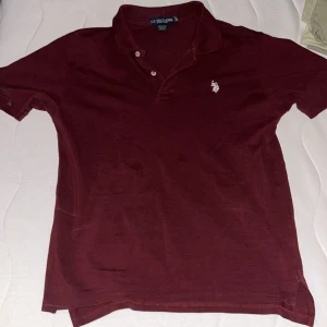 Vinröd pikétröja från U.S. Polo Assn. - Säljer en stilren vinröd pikétröja från U.S. Polo Assn. med klassisk krage och två knappar. Tröjan har en broderad logotyp på bröstet och är perfekt för en avslappnad look.