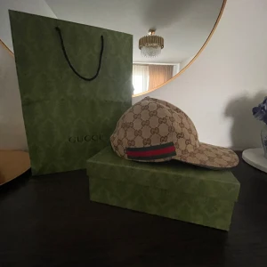 Beige keps från Gucci - Säljer en stilren beige keps från Gucci med det ikoniska GG-mönstret. Kepsen har en klassisk design med en röd och grön randdetalj. Perfekt för att ge din outfit en lyxig touch.
