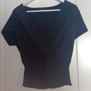 Svart omlottopp från Sisters Point - Snygg svart omlottopp från Sisters Point med korta ärmar och smockdetaljer i midjan. Perfekt för en stilren look. 🖤