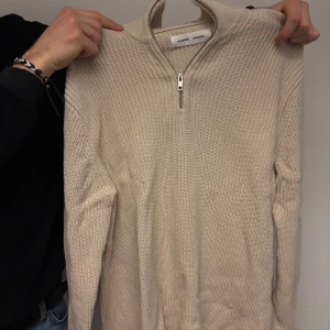 Beige stickad tröja från Samsøe Samsøe - Säljer en stilren beige stickad tröja från Samsøe Samsøe med dragkedja i halsen. Perfekt för kyliga dagar med sin mjuka och bekväma passform. Ett måste i garderoben för den som gillar klassisk stil.