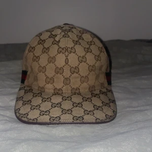 Beige keps från Gucci - Snygg beige keps från Gucci med det ikoniska GG-mönstret. Kepsen har gröna och röda ränder på sidan och är tillverkad i Italien. Perfekt för att ge din outfit en lyxig touch.