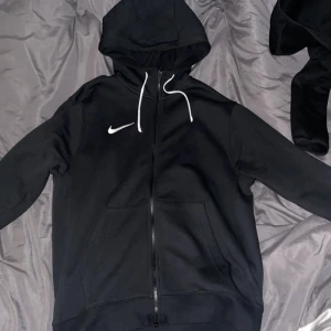 Svart hoodie från Nike - Säljer en svart hoodie från Nike med dragkedja och vit snörning i huvan. Den har en klassisk design med långärmade ärmar och en liten Nike-logga på bröstet. Perfekt för en avslappnad stil.