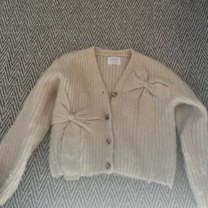 Beige stickad kofta från Zara - Säljer en mysig beige stickad kofta från Zara med dekorativa rosetter framtill. Koftan har en v-ringad design och knappar längs framsidan. 