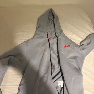 Grå tjocktröja från Hugo - Säljer en stilren grå hoodie från Hugo med röd logga på bröstet. Den har en dragkedja framtill och en praktisk huva. Perfekt för en avslappnad look. Priset kan diskuteras vid en snabbaffär och är även öppen för byten!