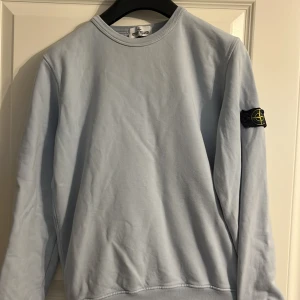 Ljusblå tröja från Stone Island - Säljer en stilren ljusblå tröja från Stone Island med deras ikoniska märke på ärmen. Tröjan har en rund hals och långa ärmar, perfekt för en avslappnad look. Passar bra till både jeans och chinos. 