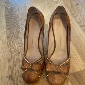 Bruna pumps från Giuseppe Zanotti - Snygga bruna pumps från Giuseppe Zanotti med krokodilmönster och klack. Skorna har en dekorativ rosett med nitar framtill och en öppen tå. Perfekta för att ge en elegant touch till din outfit.