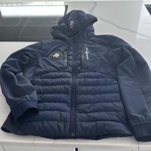 Blå pufferjacka från Sail Racing - Säljer en blå pufferjacka från Sail Racing med huva och dragkedja. Jackan har ett broderat märke på bröstet och är perfekt för kyligare väder. Den är både stilren och funktionell.
