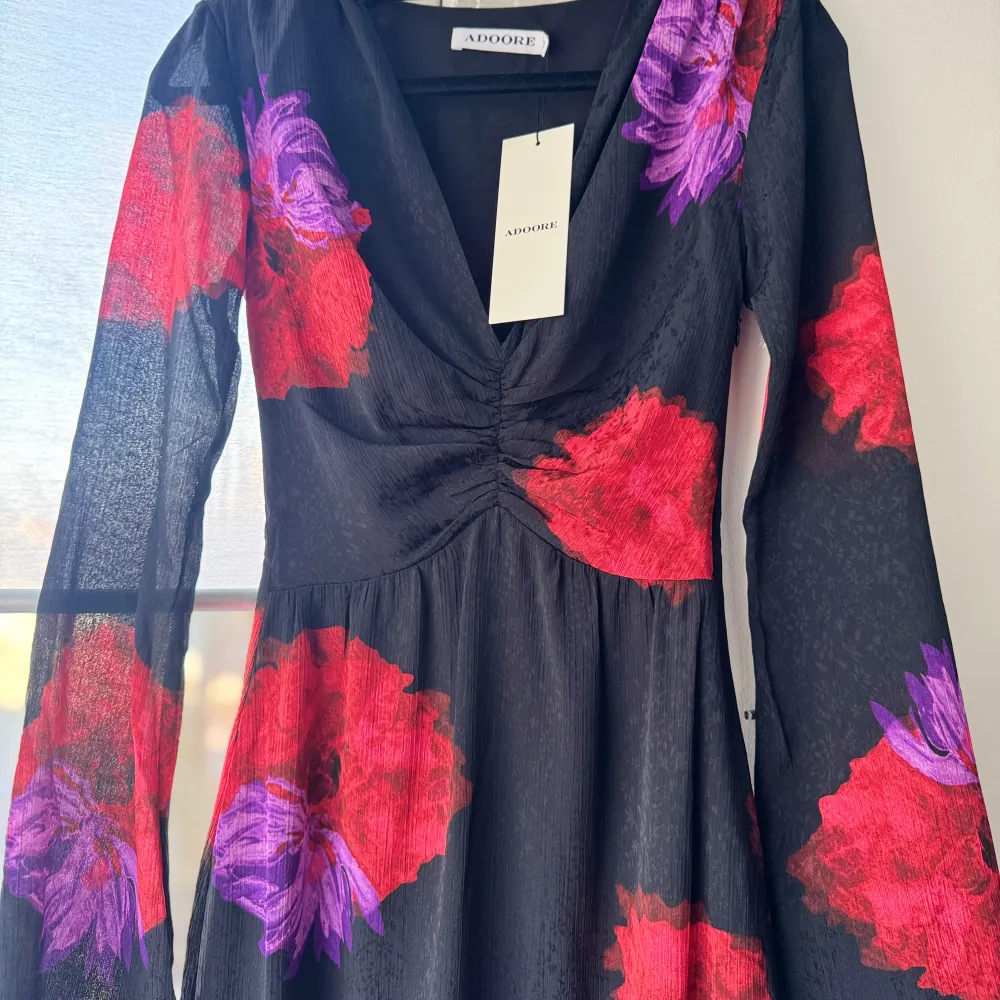 Elegant svart långklänning från ADOORE med stora röda och lila blommor. Klänningen har en v-ringad design och långa ärmar, vilket ger en stilren look. Perfekt för speciella tillfällen. Helt ny, lappar kvar. Slutsåld på hemsidan❤️💜 storlek 32. Mekot.