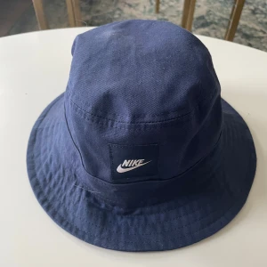 Blå fiskehatt från Nike - Snygg blå fiskehat hat från Nike med klassisk design och logga framtill. Perfekt för en avslappnad stil och skydd mot solen. Tillverkad i bomull för en bekväm passform.
