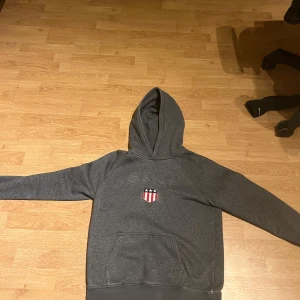 Grå hoodie från GANT - Säljer en stilren grå hoodie från GANT med broderad logga och emblem på bröstet. Tröjan har en klassisk känguruficka och en bekväm huva. Perfekt för en avslappnad stil.