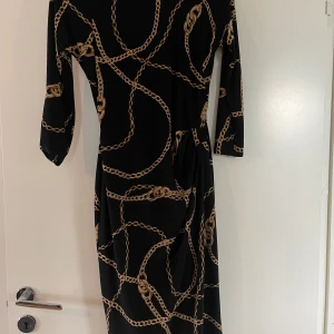 Svart långklänning med kedjemönster från Lauren Ralph Lauren - Säljer en svart vintage midiklänning från gamla kollektionen Lauren Ralph Lauren med guldiga kedjor som mönster. Klänningen har trekvartsärm och en elegant, figurnära passform. Perfekt för dig som vill sticka ut med en unik och trendig look. Säljes inte längre. 