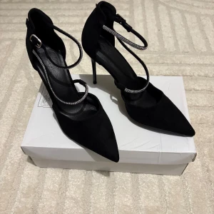 Svarta pumps med smyckade remmar - Säljer ett par eleganta svarta pumps med spetsig tå och höga klackar. Skorna har dubbla remmar, varav en är dekorerad med glittrande detaljer. Perfekta för att lyfta din outfit till nästa nivå.