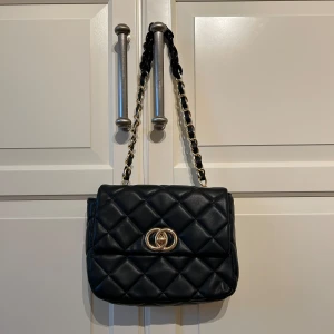 Svart quiltad axelväska från Sixth Sense - Säljer en svart quiltad axelväska från Sixth Sense med guldfärgad kedja och dekorativt spänne framtill. Väskan har en klassisk design med lock och kedjerem som är flätad med svart material. Perfekt för att lyfta din outfit.