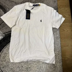 Vit t-shirt från Polo Ralph Lauren - Klassisk vit t-shirt från Polo Ralph Lauren med rund halsringning och liten broderad logga på bröstet. Tillverkad i mjuk bomull för en skön känsla. Perfekt basplagg med enkel och stilren design.