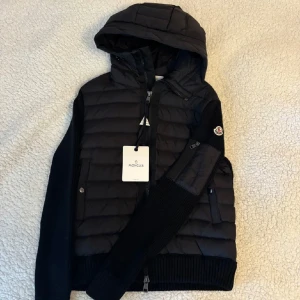 Moncler Hooded Cardigan – helt ny & stilren - Svart zip-up hoodie i äkta Moncler-kvalitet. Mjuk stickad känsla, lätt vadderade detaljer och huva – perfekt för både vardag och mer dressad stil. Ikoniska loggan på ärmen ger en tydlig men clean look. Aldrig använd – nypris runt 6 000 kr. Du får den helt ny, till ett mycket bättre pris. Bara en tillgänglig – först till kvarn.