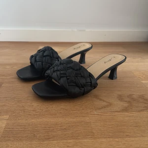 Svarta flätade klack-sandaler från Alley - Säljer ett par oanvända svarta sandaler med klack från Alley. Skorna har en bred, flätad rem över foten och fyrkantig tå. Perfekta för dig som gillar stilrena och trendiga skor med en twist. Klacken är medelhög och ger en snygg siluett.
