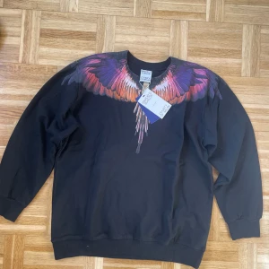 Svart sweatshirt med färgglatt vingmönster från Marcelo Burlon - Säljer en svart sweatshirt från Marcelo Burlon med ett stort färgglatt vingmönster i lila, rosa och orange över axlar och bröst. Tröjan har rund hals och långa ärmar. Perfekt statement-plagg för dig som gillar unika detaljer.