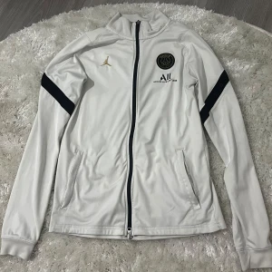 Vit träningsjacka från Jordan x PSG - Säljer en äkta  vit träningsjacka från Jordan x PSG med svarta detaljer på ärmarna och dragkedja framtill. Jackan har två fickor med dragkedja och klubbmärke på bröstet. Perfekt för fotbollsträning eller sportig stil. Köpt för ett par år sedan men nästan aldrig kommit till användning.