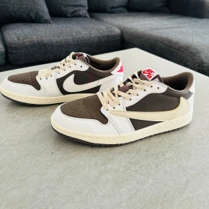 Nike Air Jordan 1 Low Travis Scott x Fragment - Säljer ett par Nike Air Jordan 1 Low Travis Scott x Fragment med bruna, vita och beige paneler, ikonisk omvänd swoosh och röda detaljer. Skorna har snörning och platt sula, perfekt för dig som gillar streetwear och sneakerskultur.