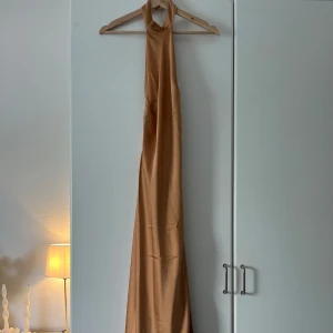 Champagnefärgad långklänning från Lovisa Barkman x NA-KD - Säljer en elegant långklänning i satin från Lovisa Barkman x NA-KD. Klänningen är champagnefärgad med öppen rygg och halterneck. Materialet är glansigt och mjukt, perfekt för speciella tillfällen.🧡