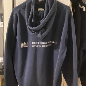 Mörkblå hoodie från Drottning Blankas Gymnasieskola - Mörkblå hoodie med tryck från Drottning Blankas Gymnasieskola. Tröjan har huva med dragsko och en stor magficka framtill. Perfekt för en avslappnad stil.