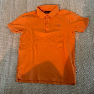 Orange pikétröja från Tommy Hilfiger - Säljer en orange pikétröja från Tommy Hilfiger med klassisk krage och knappar framtill. Liten broderad logga på bröstet. Perfekt för en stilren och sportig look.