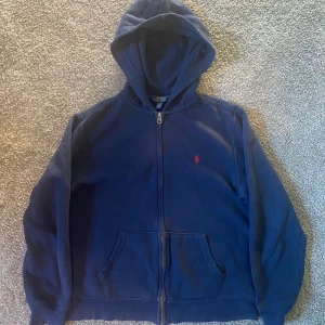 Mörkblå hoodie från Polo Ralph Lauren - Mörkblå hoodie från Polo Ralph Lauren med dragkedja och huva. Klassisk röd broderad logga på bröstet och två fickor framtill. Perfekt för en avslappnad stil och enkel att matcha med det mesta.