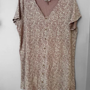 Rosa blommig klänning med knappar från Divided - Söt rosa kortklänning från Divided med vitt blommönster och v-ringning. Klänningen har korta ärmar och knappar framtill, vilket ger en avslappnad och feminin look. Perfekt för varma dagar och enkel att styla med sneakers eller sandaler.