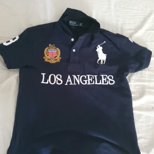 Mörkblå pikétröja från Polo Ralph Lauren - Säljer en mörkblå pikétröja från Polo Ralph Lauren med vit Los Angeles-tryck på framsidan, stor vit logga och broderad emblem. Nummer 3 på ryggen och ärmarna. Klassisk krage och knappar framtill.