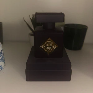 Initio side effect 90ml - Elegant parfymflaska i matt lila med fyrkantig form och en stor, fyrkantig kork. Flaskan har en dekorativ guldfärgad detalj i form av en kvadrat på framsidan. Perfekt som snygg inredningsdetalj eller till din doftsamling.