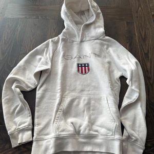 Vit hoodie från GANT - Säljer en vit hoodie från GANT med broderad logga och sköld med stjärnor och ränder på bröstet. Tröjan har huva och en stor magficka framtill. Hoodien är i fint skick utan några defekter. Skriv till mig om ni har några frågor!