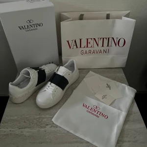 Valentino  skor - Säljer ett par snygga vita sneakers från Valentino Garavani med ett brett svart band över mitten. Skorna har rund tå, platt sula och klassisk snörning. Kommer med originalkartong, dustbag och påse.