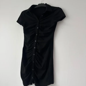 Svart kortärmad skjortklänning med knappar - Säljer en svart skjortklänning med korta ärmar och knappar längs hela framsidan. Klänningen har en krage och lätt rynkade detaljer längs knappraden för en snygg passform. Perfekt för dig som gillar stilrena och enkla plagg.