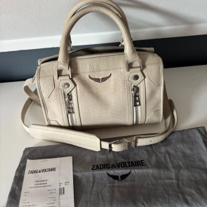 Beige handväska från Zadig & Voltaire - Snygg beige handväska från Zadig & Voltaire i skinn med ormmönstrad struktur. Väskan har dubbla handtag, avtagbar axelrem och två dekorativa dragkedjor framtill. Nästan aldrig använd. Nypris: 6100kr 