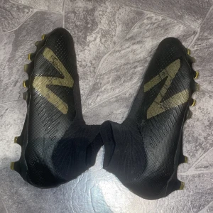 Svarta fotbollsskor från New Balance med guldiga detaljer!PRIS KAN DISKUTERAS  - Säljer ett par svarta fotbollsskor från New Balance med stora guldiga N-loggor på sidorna. Skorna har snörningslös design, högre strumpliknande skaft och fasta dobbar för spel på gräs. Perfekta för dig som vill sticka ut på planen. Även signerade av en före detta Allsvenskan spelare