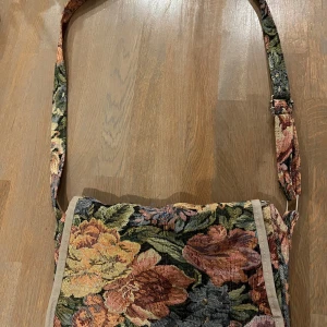 Blommig axelväska i gobelängtyg - Säljer en unik axelväska med blommigt gobelängmönster i grönt, blått, gult, rosa och beige. Väskan stängs med dragkedja. Perfekt för dig som vill ha en personlig accessoar med vintagekänsla.
